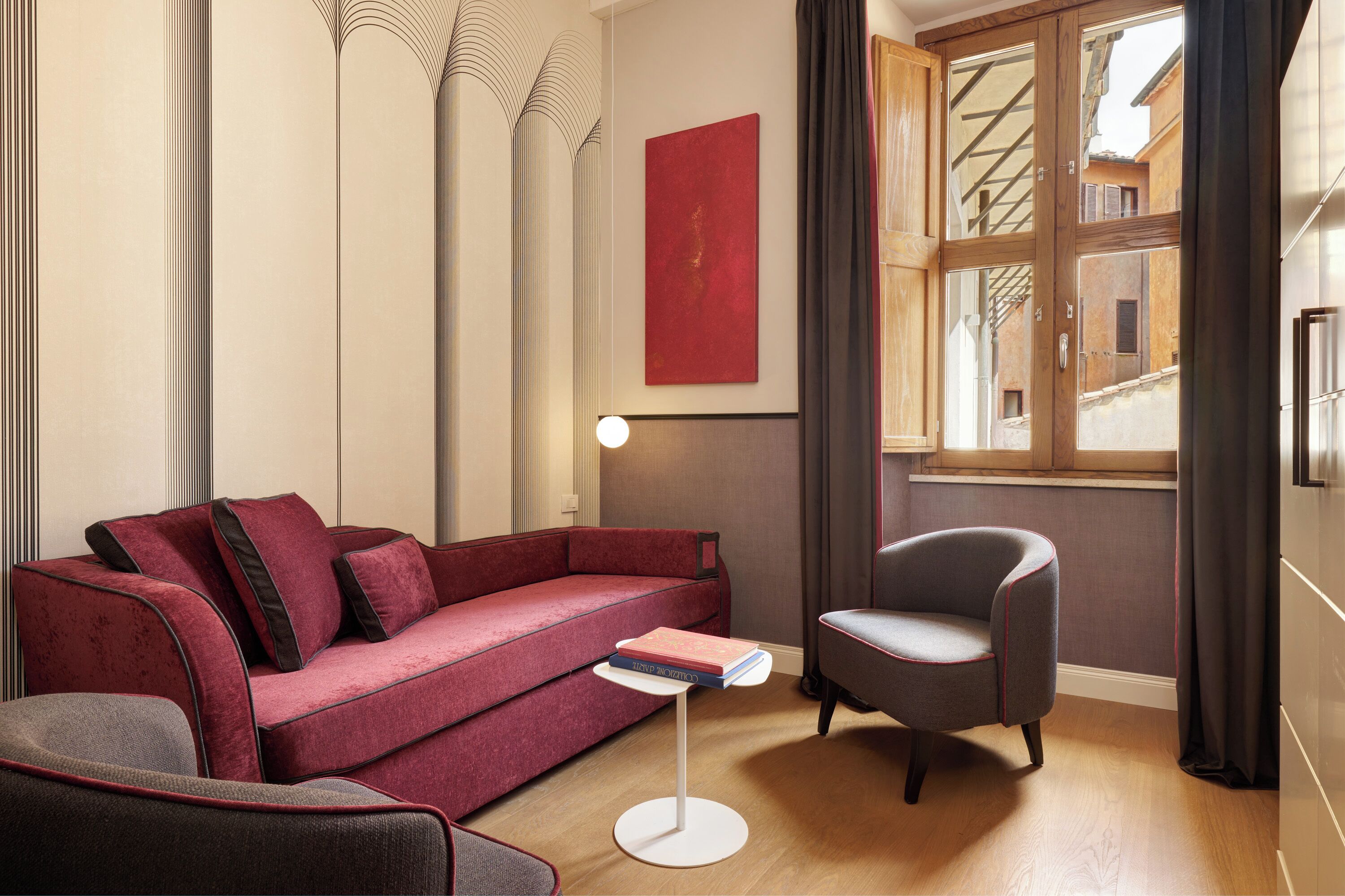 Una habitación moderna con un sofá rojo, un sillón gris, una pequeña mesa blanca y un amplio ventanal con cortinas.