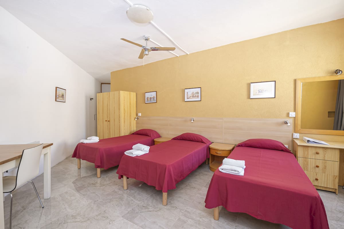 A hotel room with two single beds, a wooden desk, a chair, a wardrobe, and a ceiling fan.