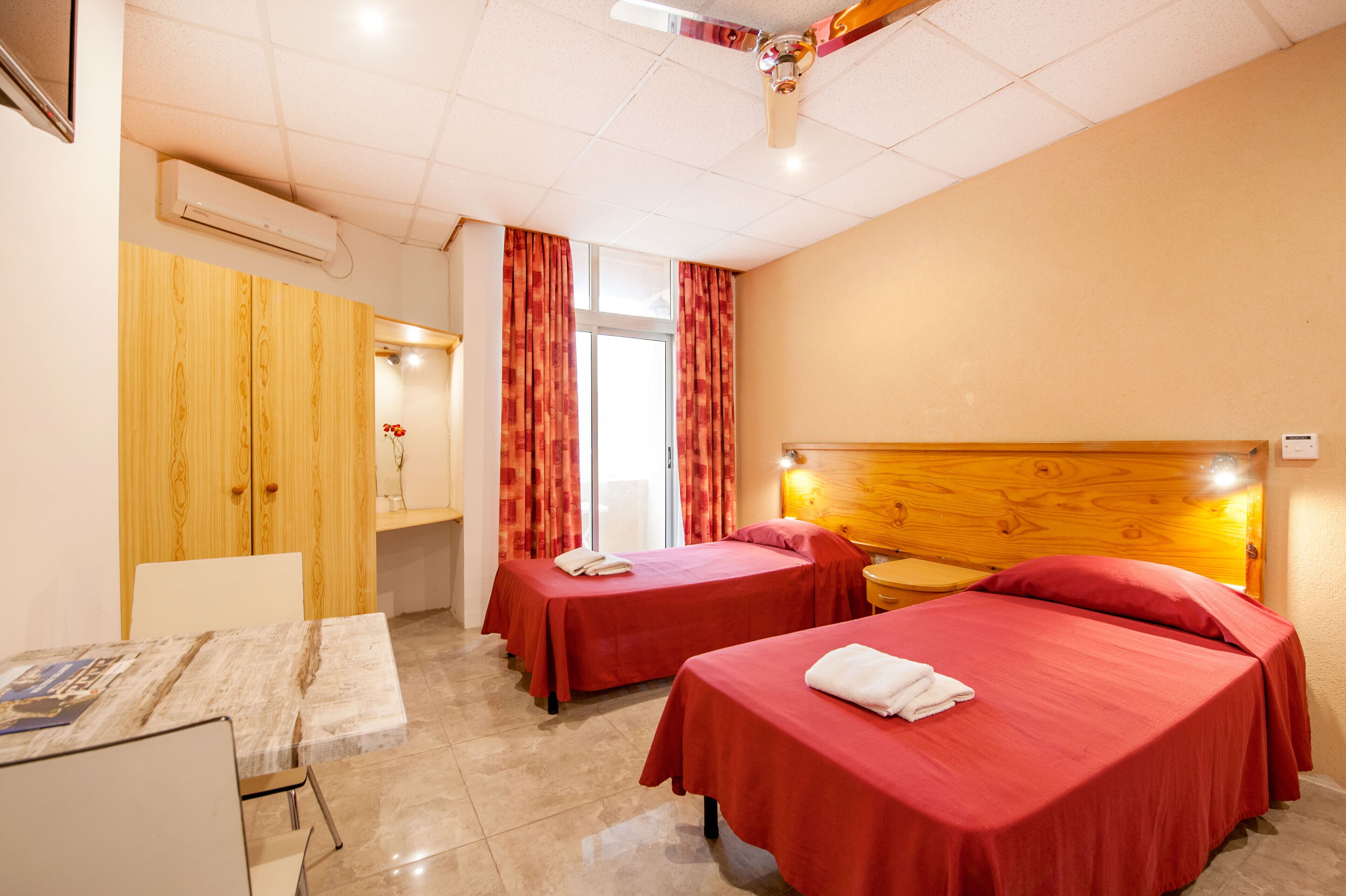 A hotel room with two single beds, a wooden wardrobe, a desk with a chair, and a window with curtains.
