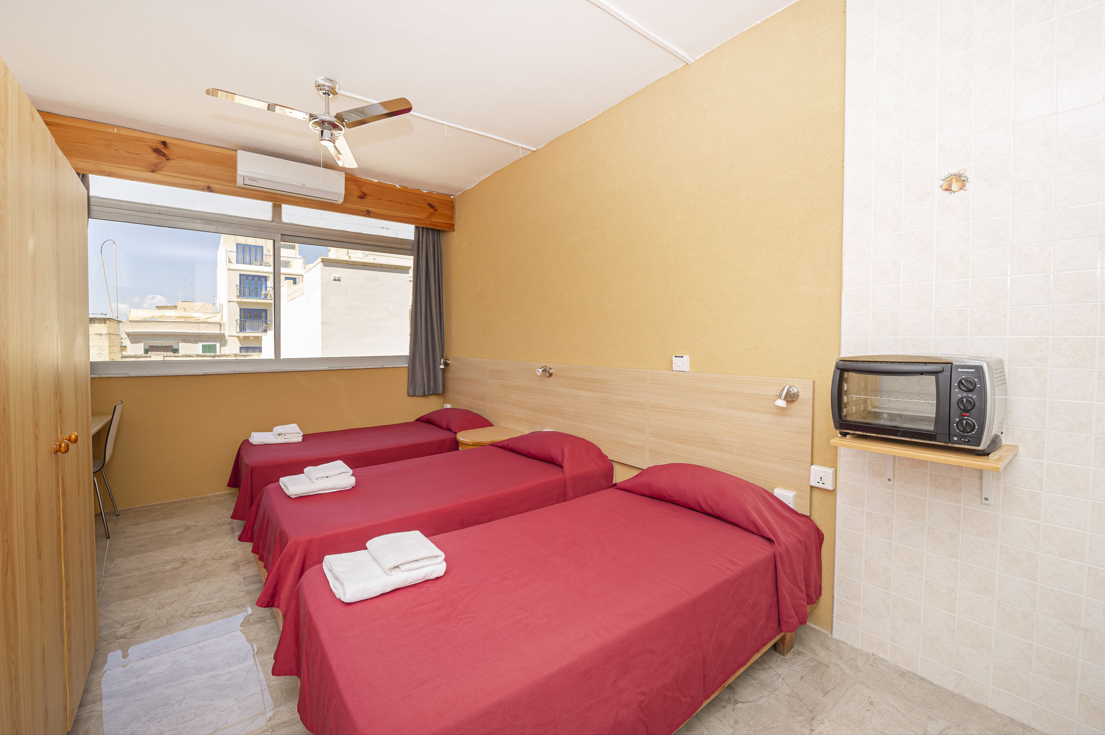 A hotel room with three single beds, a ceiling fan, a microwave, and a window with a view of buildings.