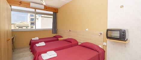 A hotel room with three single beds, a ceiling fan, a microwave, and a window with a view of buildings.