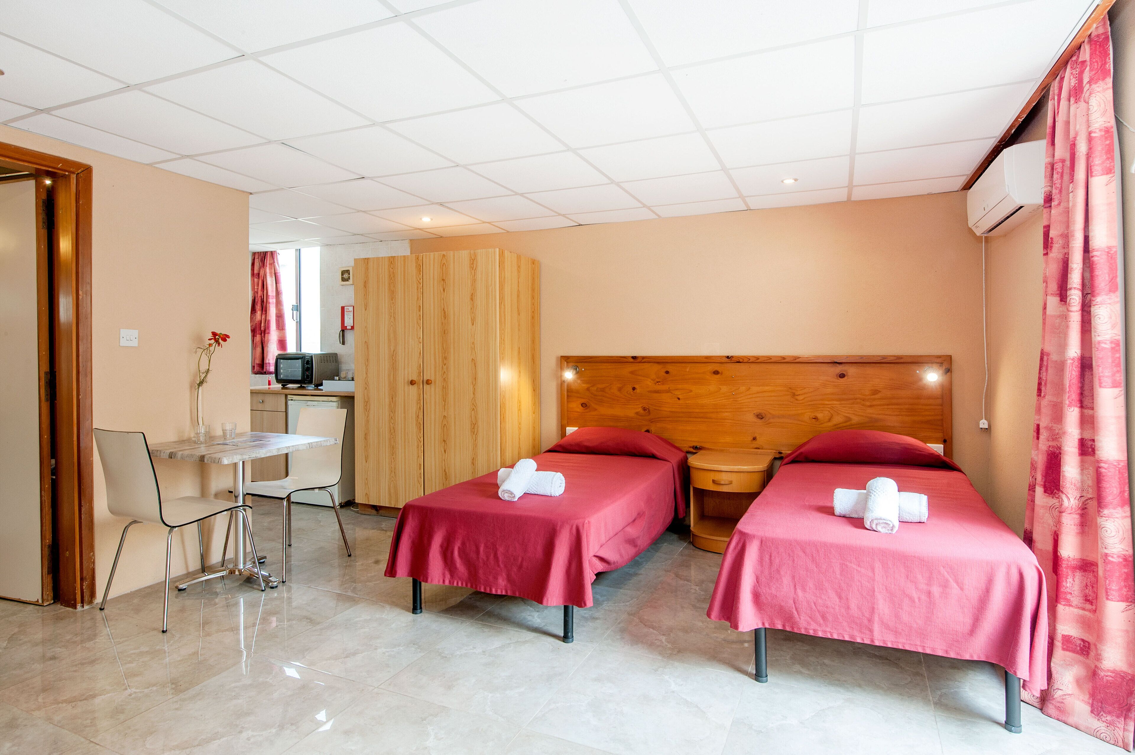 A hotel room with two single beds, a wooden headboard, a small desk with a chair, and a view of the room's interior.