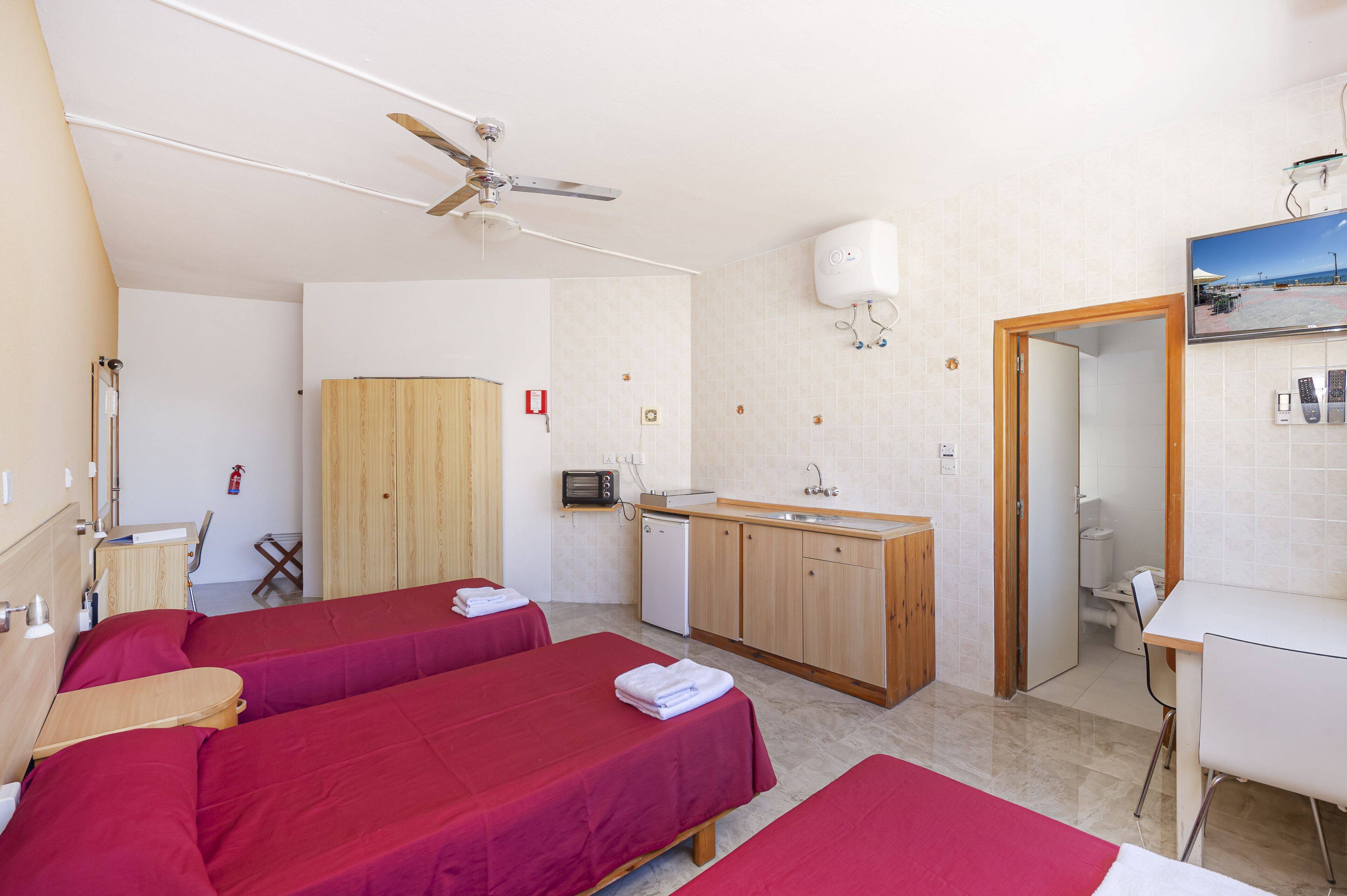 A hotel room with two beds, a kitchenette, a ceiling fan, a fire extinguisher, and a bathroom.