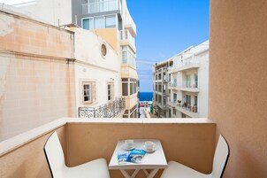 Un balcón con una mesa, dos sillas y una revista. El balcón da a una calle con edificios y vistas al mar.