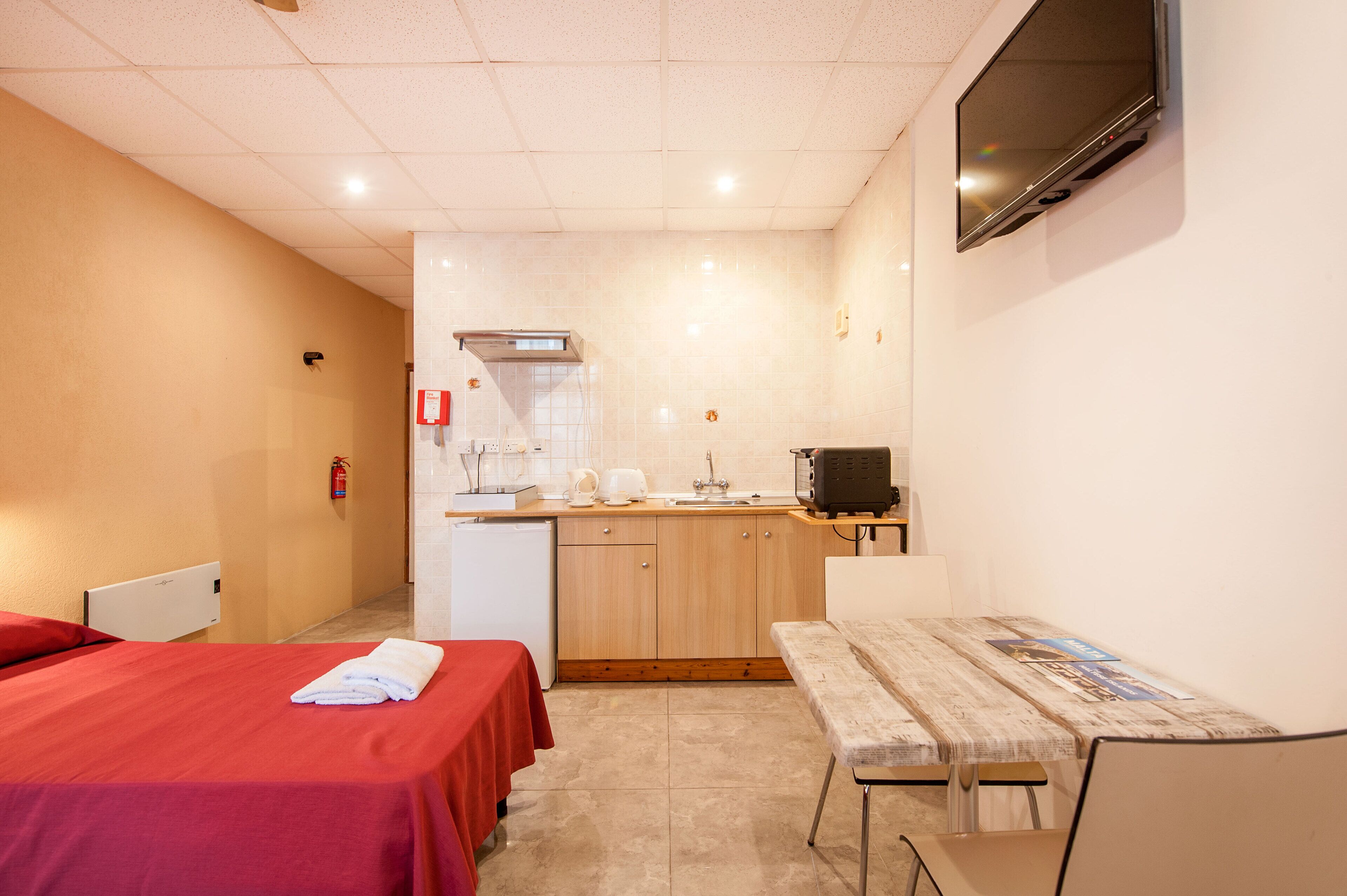 A compact hotel room with a kitchenette, a bed with a red cover, a small table, and a TV mounted on the wall.