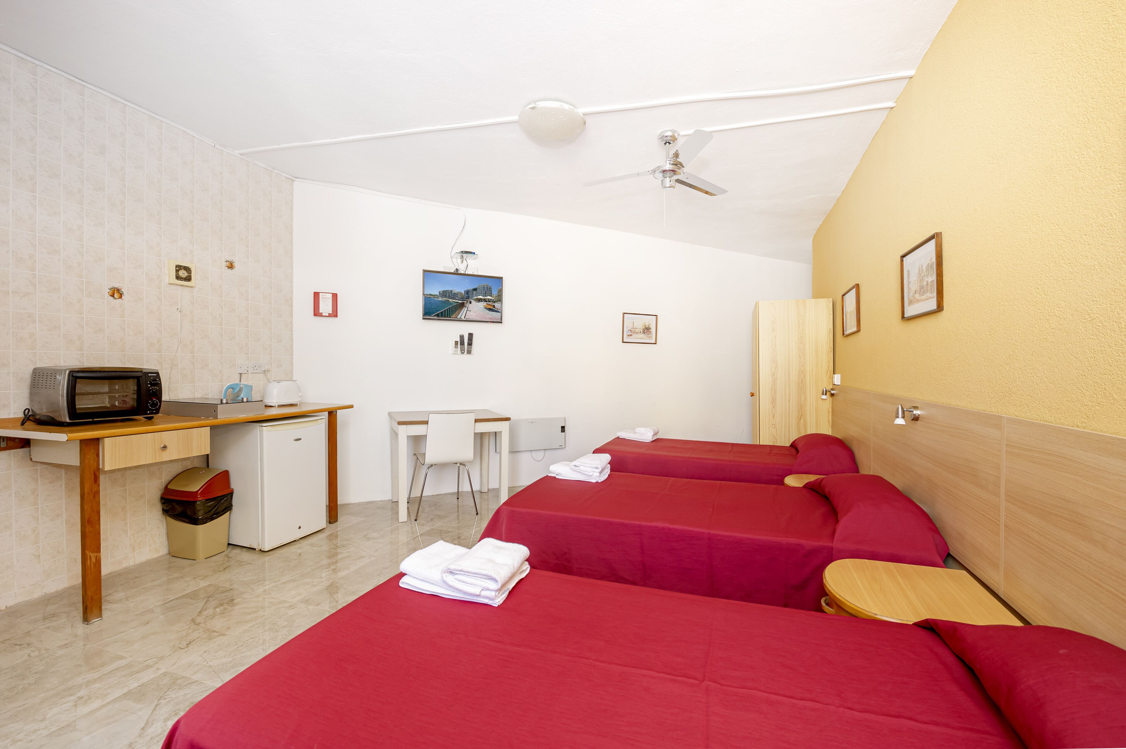 A hotel room with a desk, microwave, and a small fridge. There are three beds with red bedding and a ceiling fan.