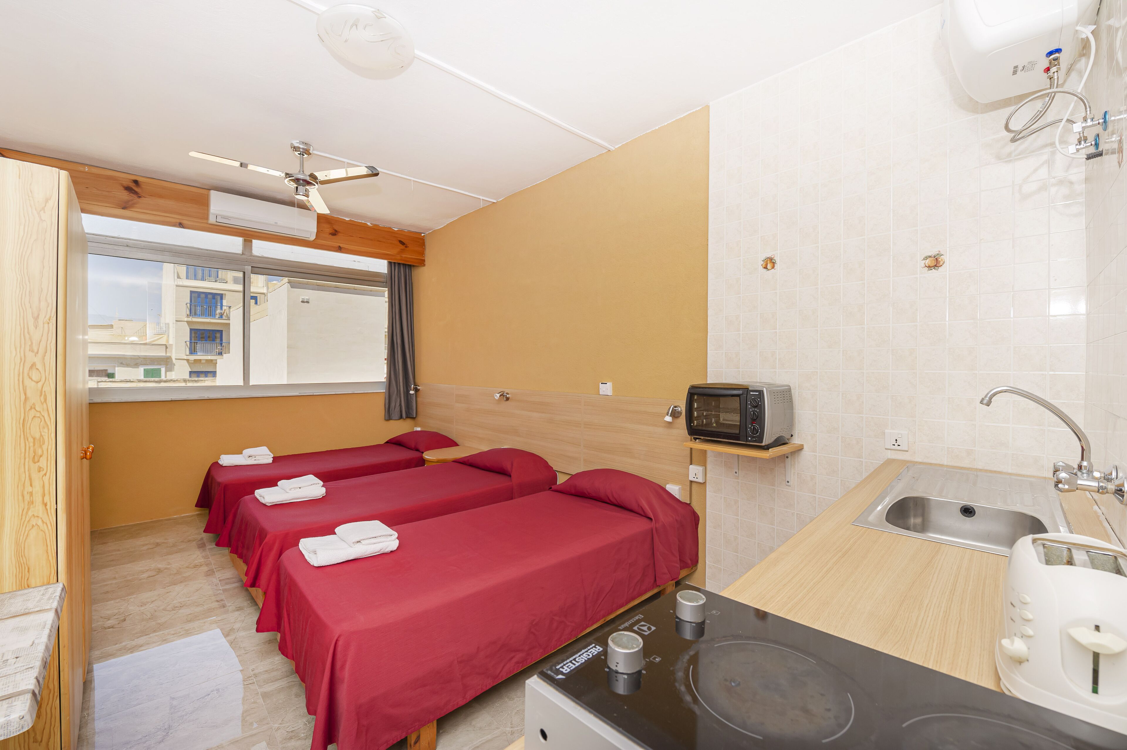 A hotel room with two single beds, a kitchenette, a microwave, a sink, and a window with a view of buildings.