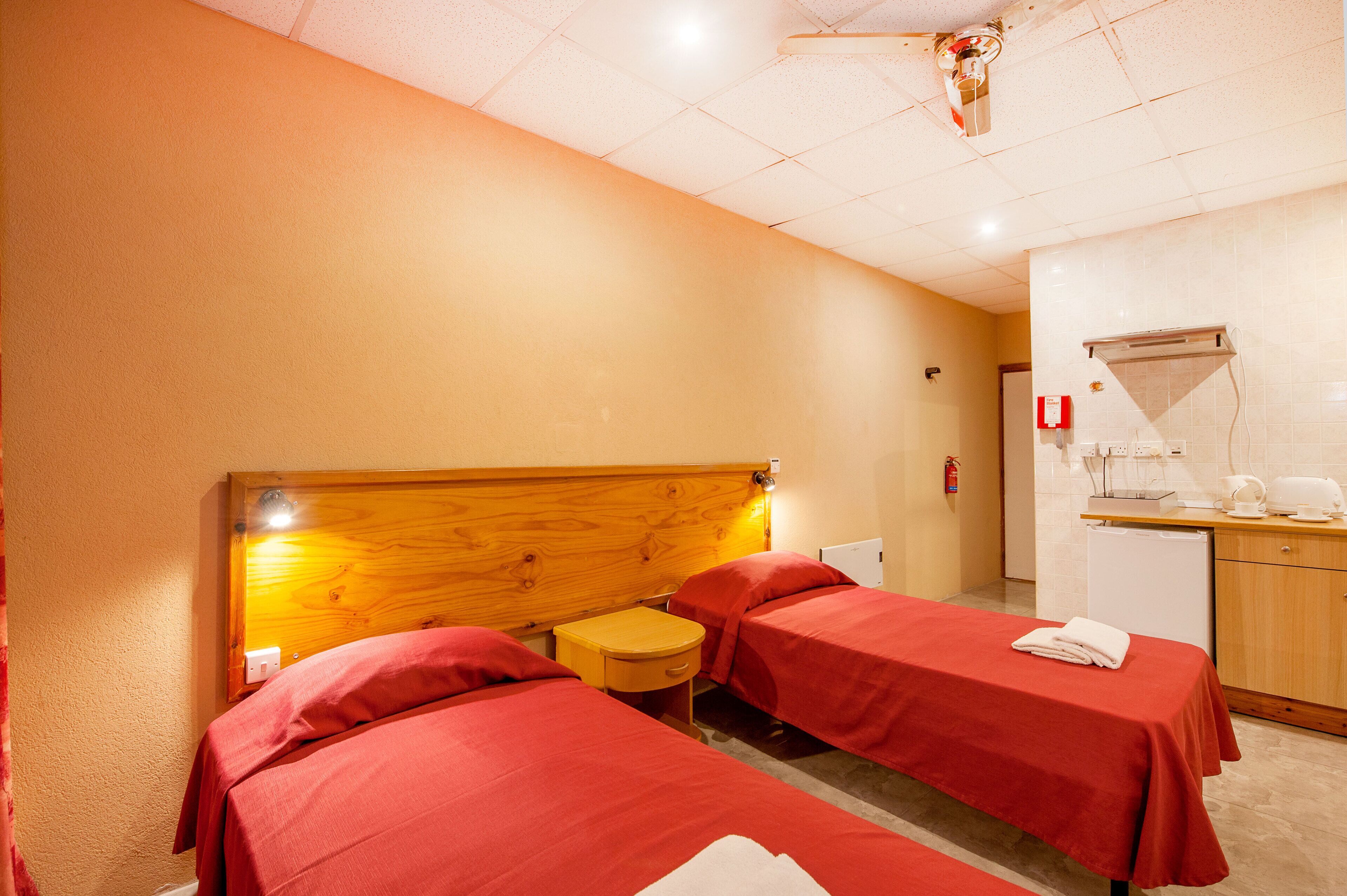 A hotel room with two single beds, a wooden headboard, a small bedside table, a red blanket, a white towel, a kitchenette with a microwave, and a ceiling fan.