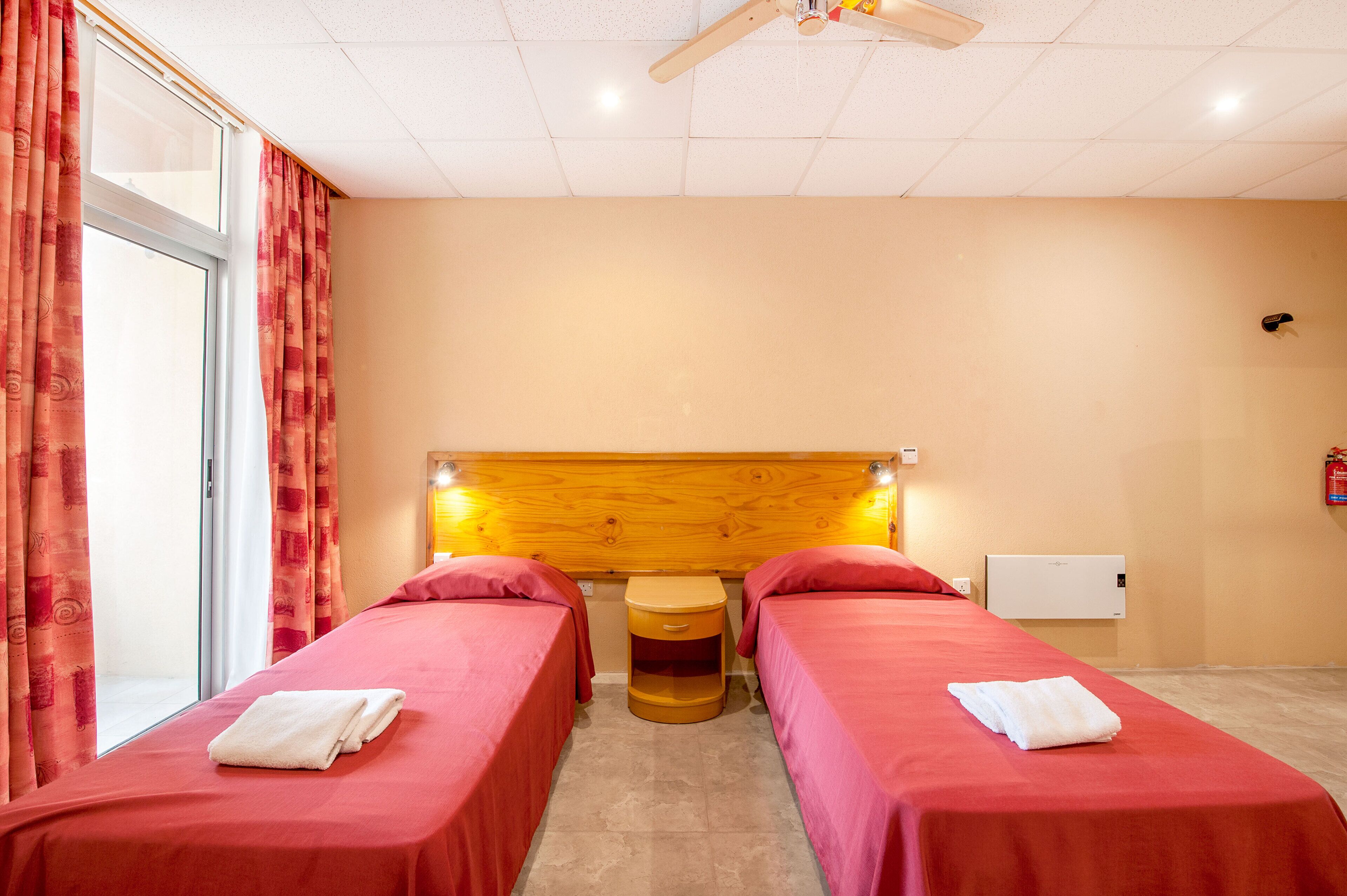A hotel room with two single beds, a wooden headboard, a small table, and a window with curtains.