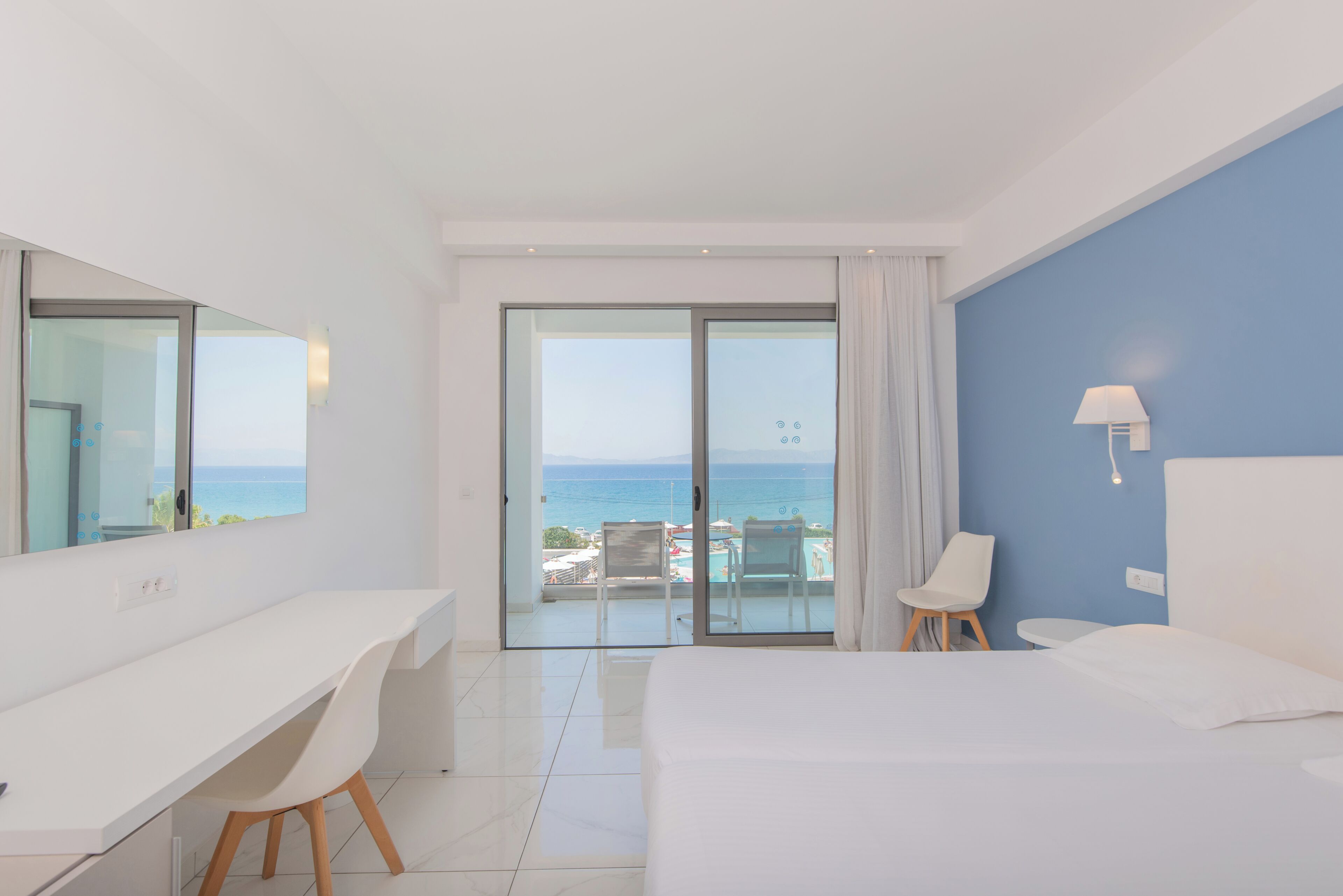 A hotel room with a bed, a desk, a chair, a small table, a lamp, and a view of the sea through a glass door.