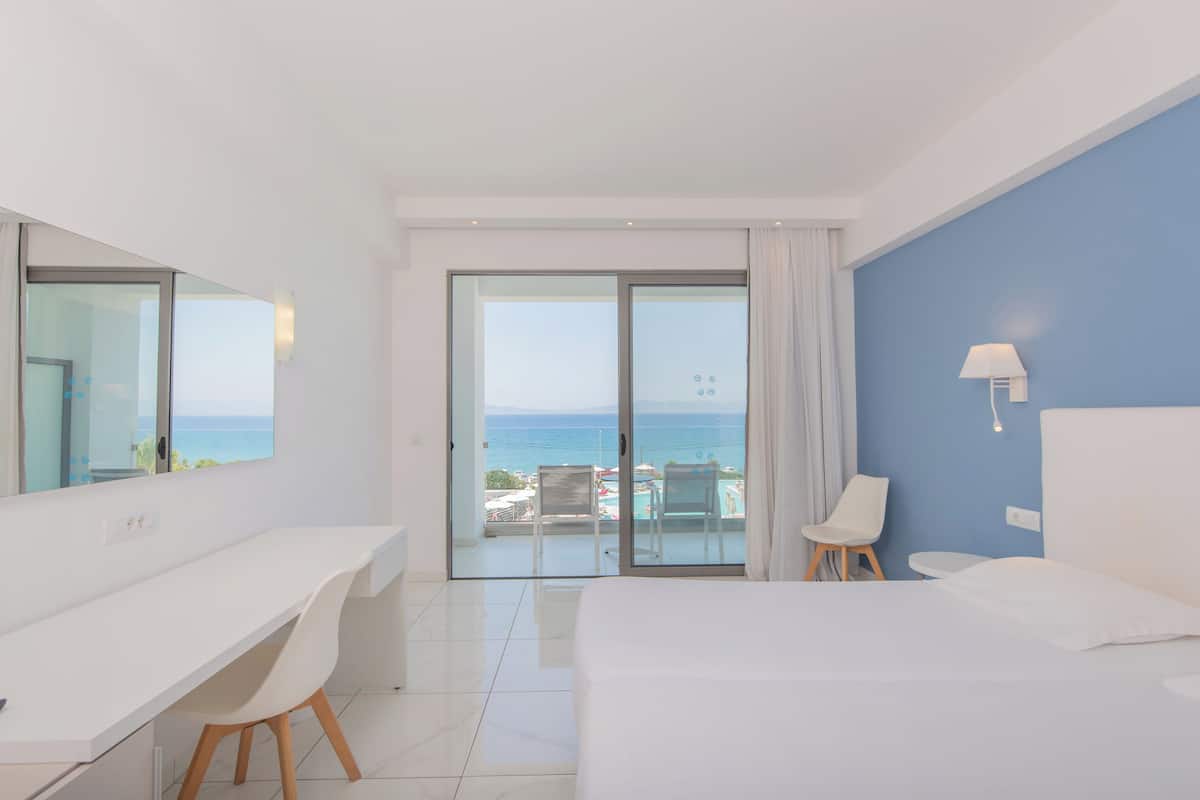 A hotel room with a bed, a desk, a chair, a small table, a lamp, and a view of the sea through a glass door.