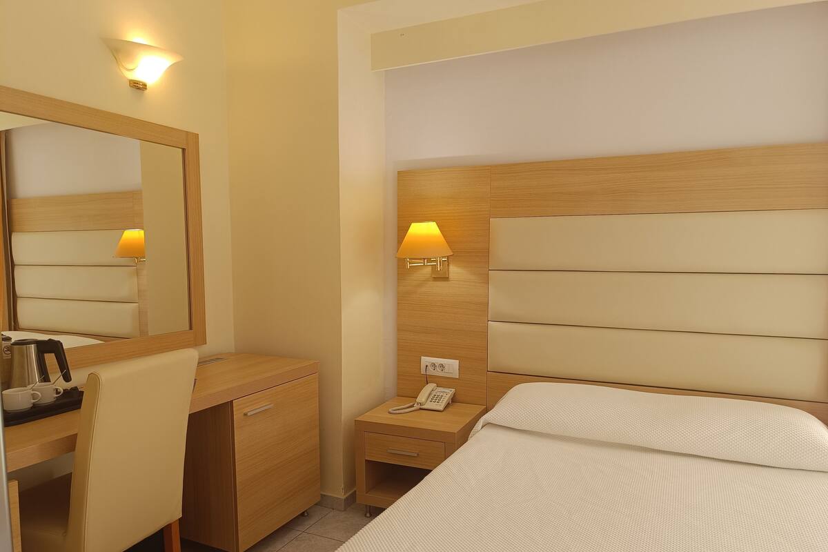 Habitación de hotel con cama, escritorio, silla, espejo y mesita de noche con lámpara y teléfono.