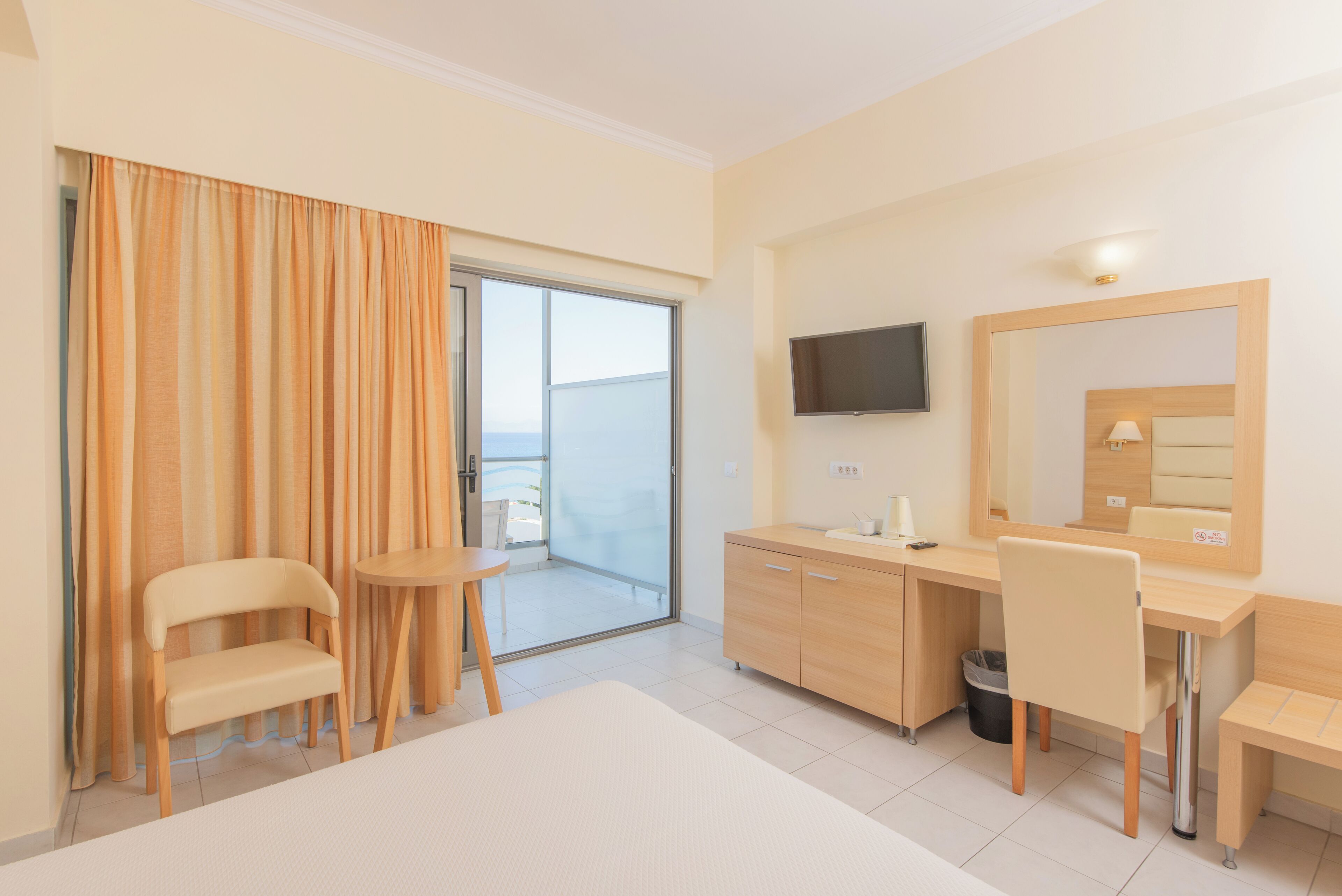 Habitación de hotel con cama, escritorio, silla, televisor, espejo y puerta corredera de cristal que da a un balcón.