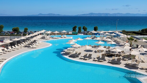 Une piscine d’un complexe hôtelier, dotée de chaises longues et de parasols, entourée d’une mer d’un bleu limpide et avec des montagnes en toile de fond.