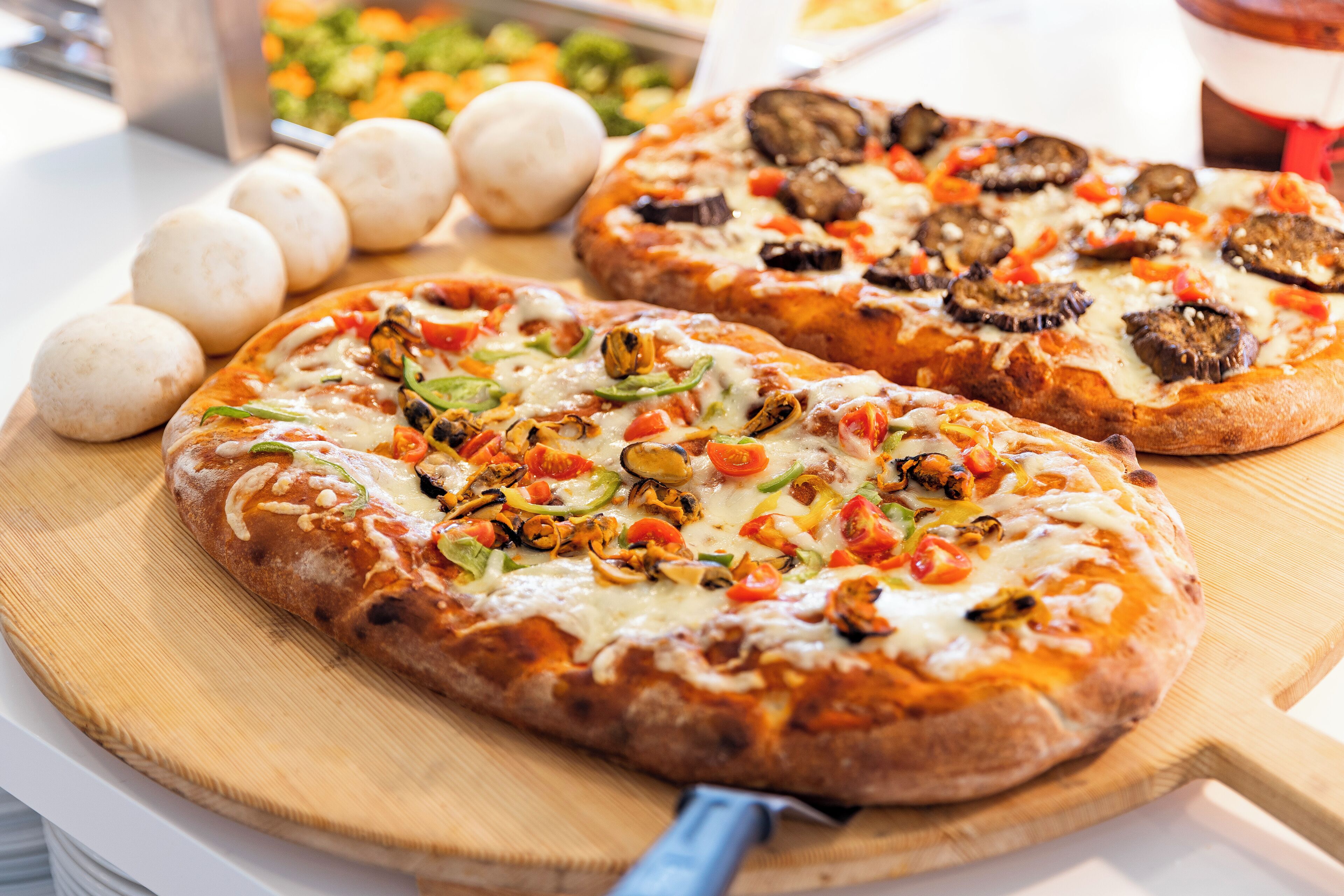 Két pizza, különféle feltétekkel, fából készült tálalódeszkán, egy pizzavágó, és a háttérben egy tál tésztagombóccal.