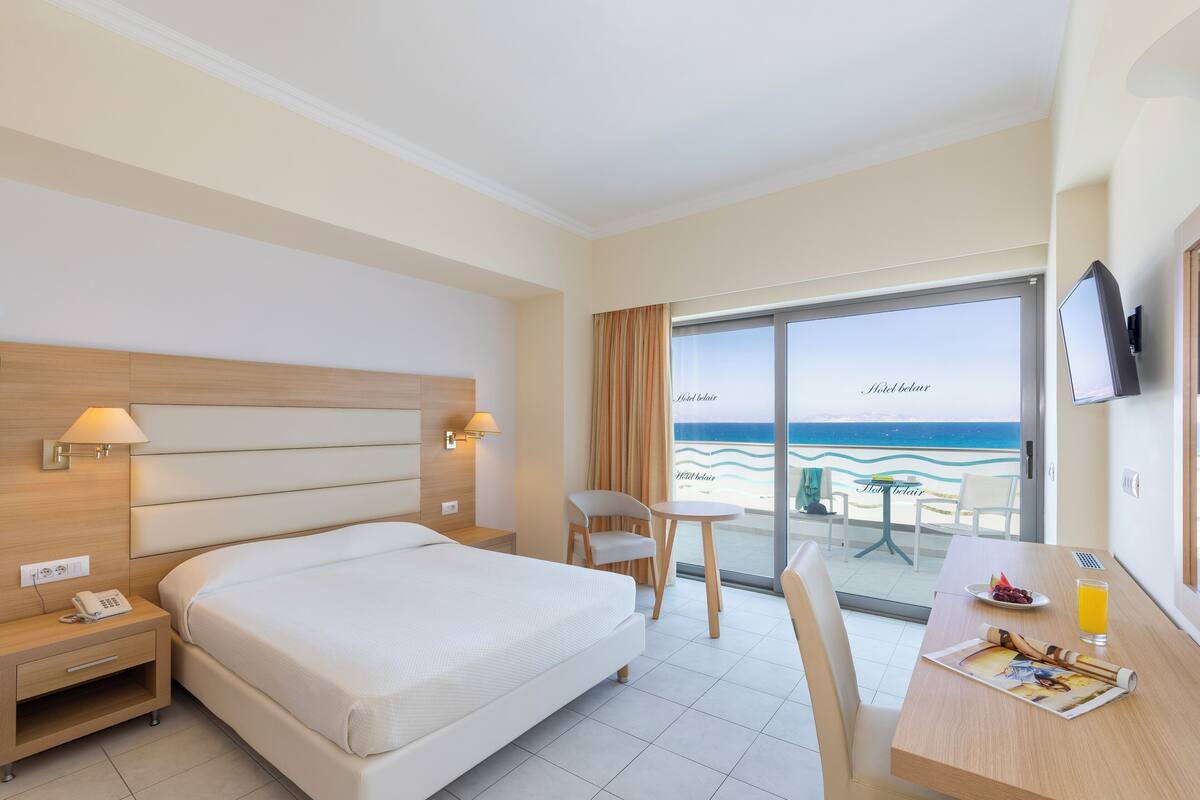 Habitación de hotel con cama, mesita de noche, lámpara, silla, mesa con desayuno y vista a la playa.