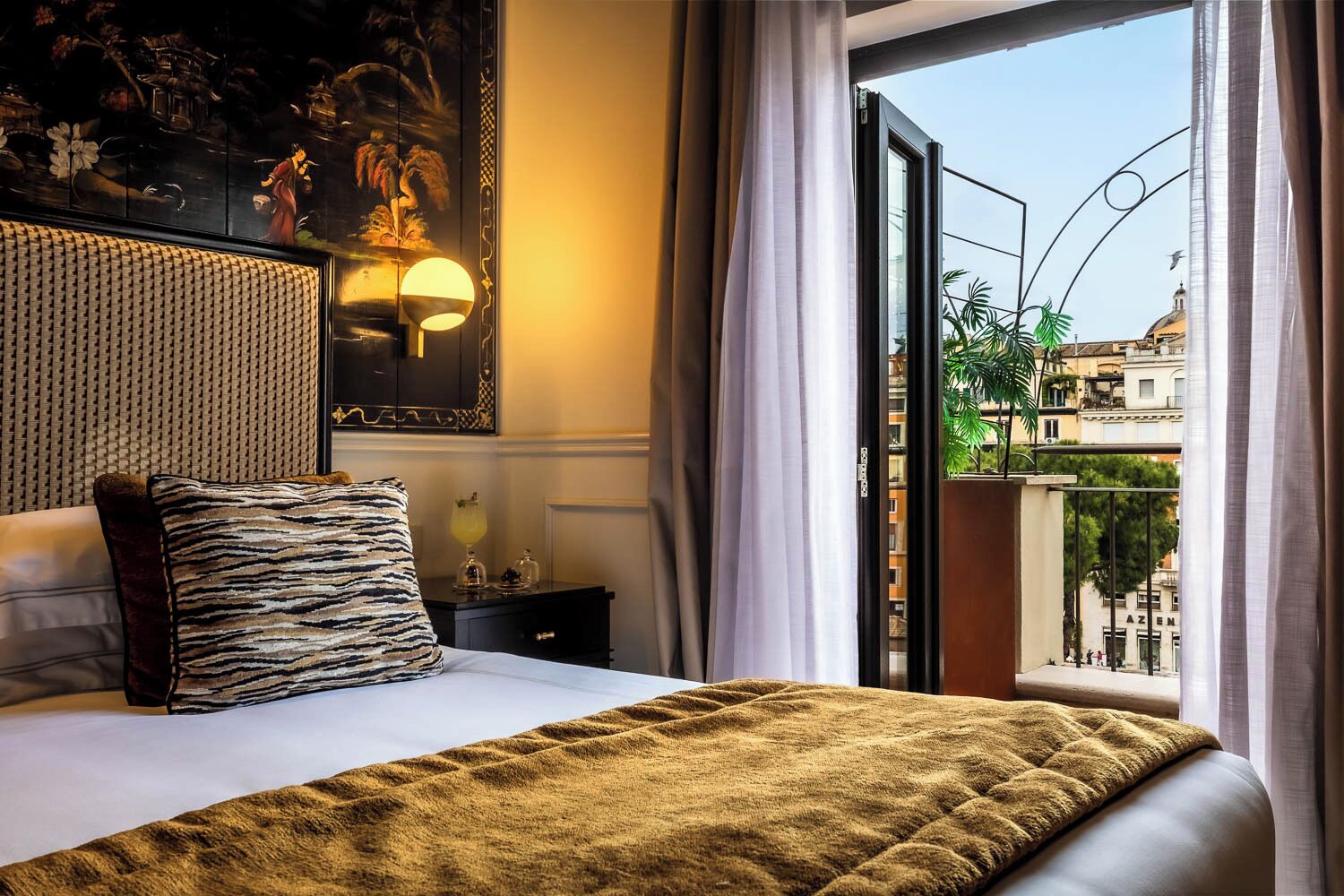 Une chambre d’hôtel avec un lit, une table de chevet, une lampe, un tableau au mur et une vue sur la ville depuis un balcon.