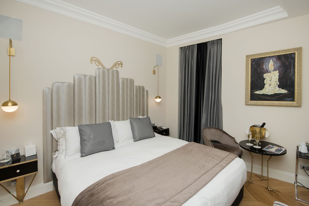 A hotel room with a large bed, a bedside table with a lamp, a chair, and a small table with a magazine and a bottle.