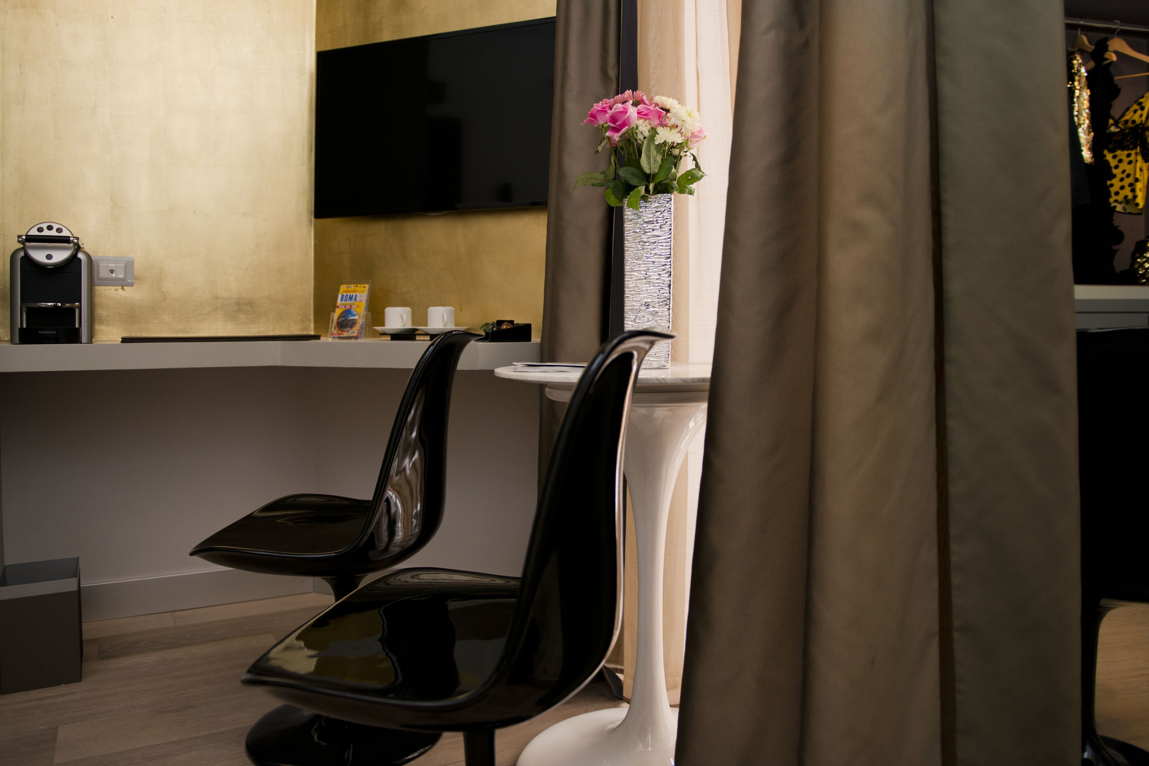 A modern hotel room with a flat-screen TV mounted on the wall, a coffee machine, a vase with flowers, and a dining area with chairs and a table.