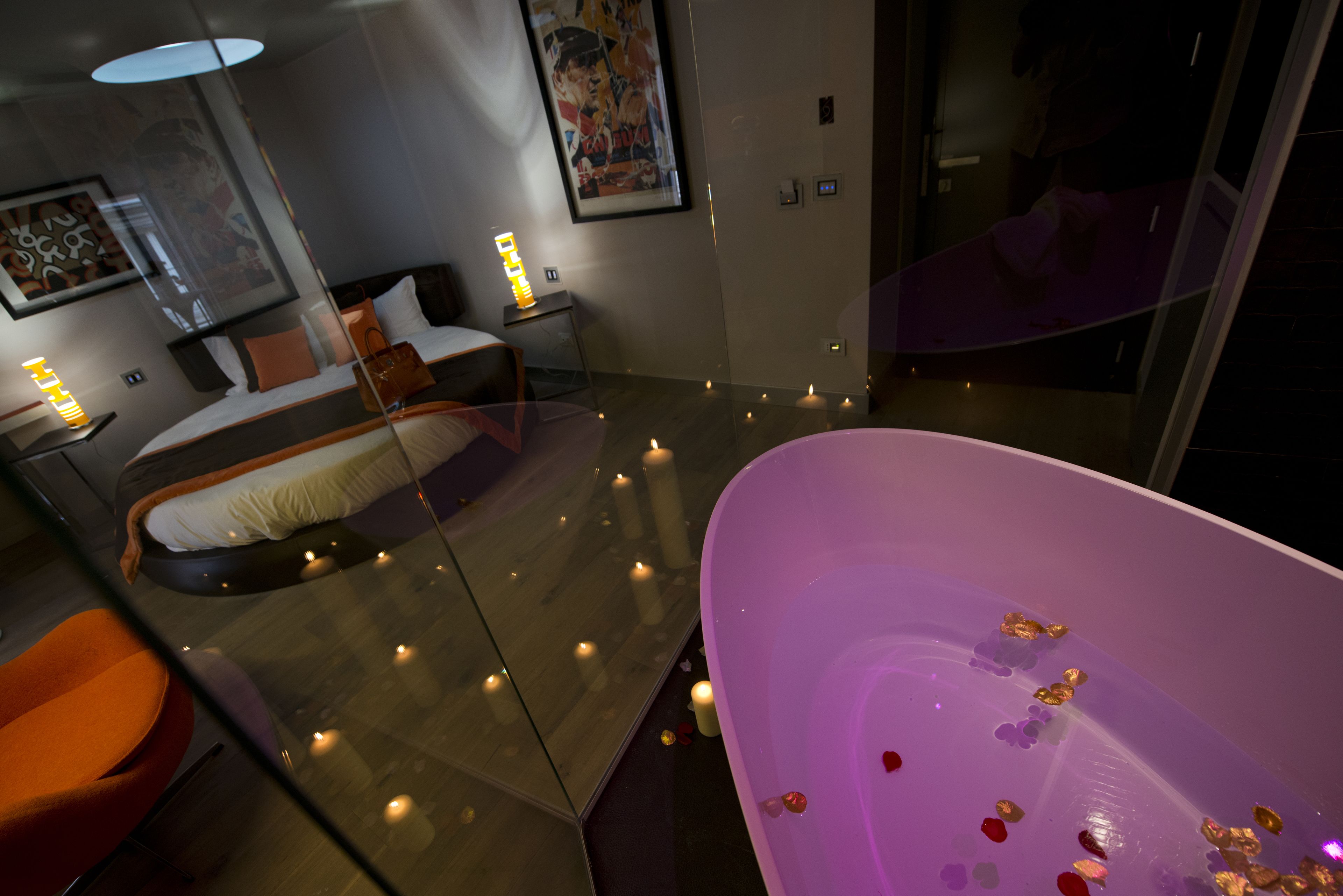 A modern hotel room with a glass-enclosed bathtub, a bed with a white and orange headboard, and a large painting on the wall.