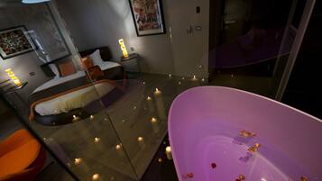 Ein modernes Hotelzimmer mit einer gläsernen Badewanne, einem Bett mit weißem und orangefarbenem Kopfteil und einem großen Gemälde an der Wand.