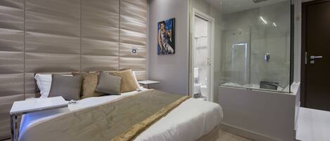 Una habitación de hotel moderna con una cama grande, una mesita de noche, un box de ducha de vidrio y un cuadro en la pared.