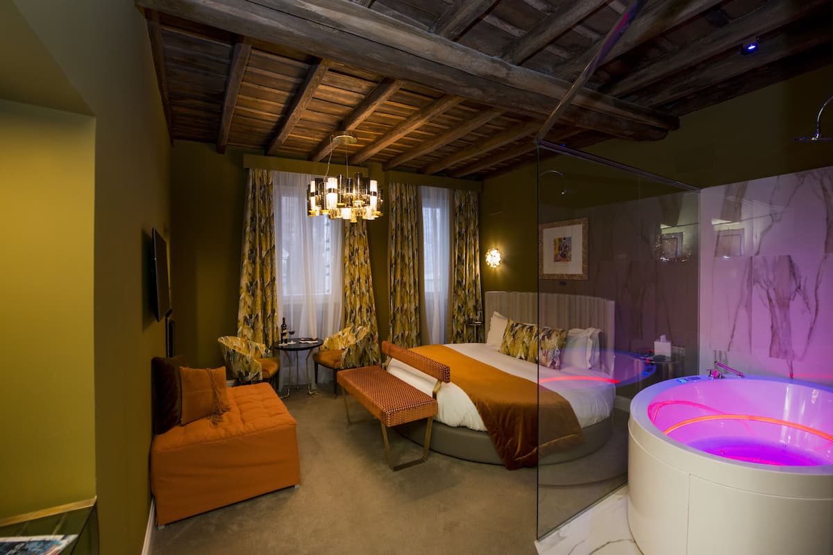 A hotel room with a bed, a bench, a small table, a large mirror, and a jacuzzi.