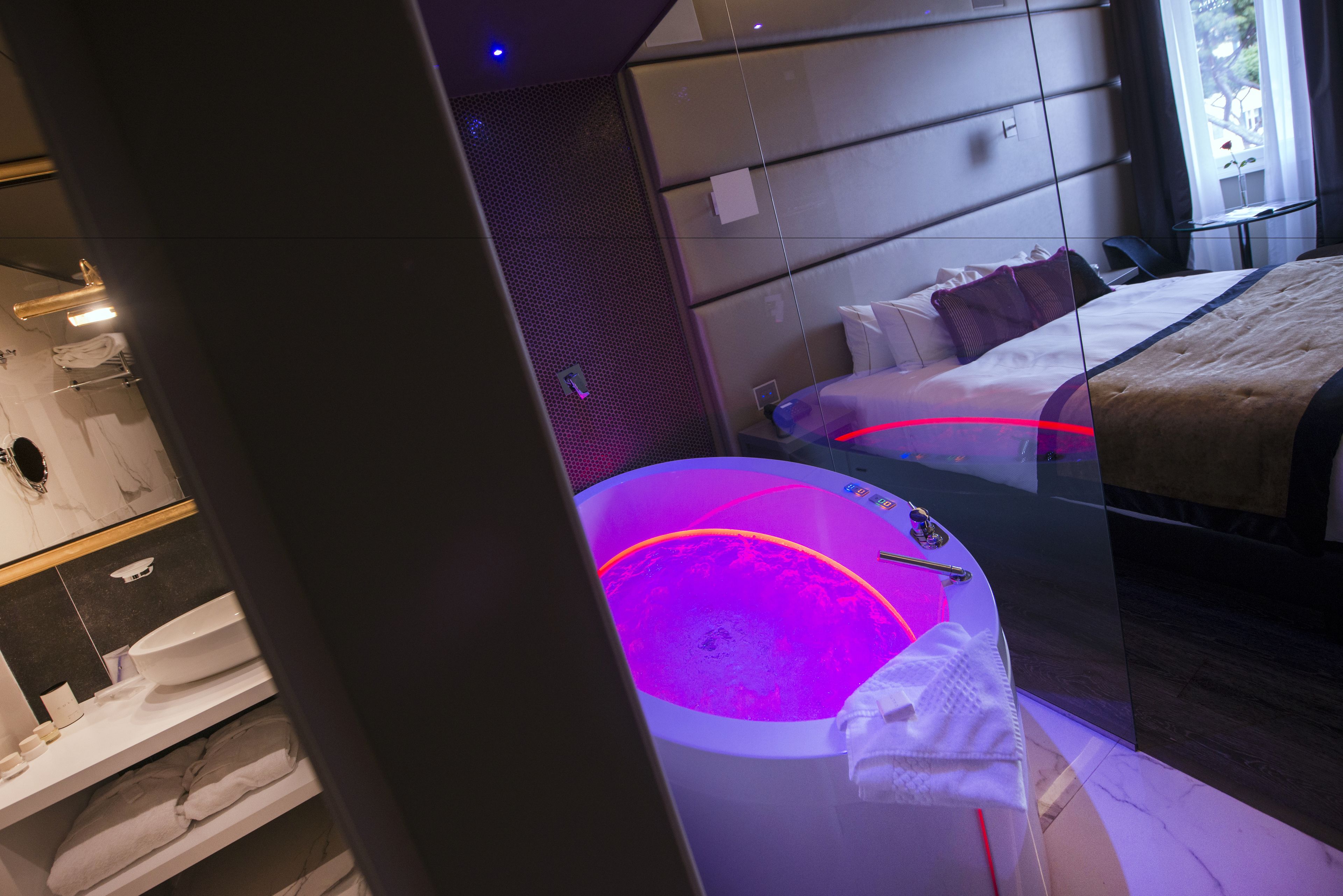 A modern hotel room with a glass partition, a bed, a bedside table, and a visible bathroom area with a bathtub.