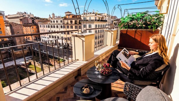 Un balcone con vista sulla città, un tavolo con bevande e frutta, e una persona che legge una rivista.