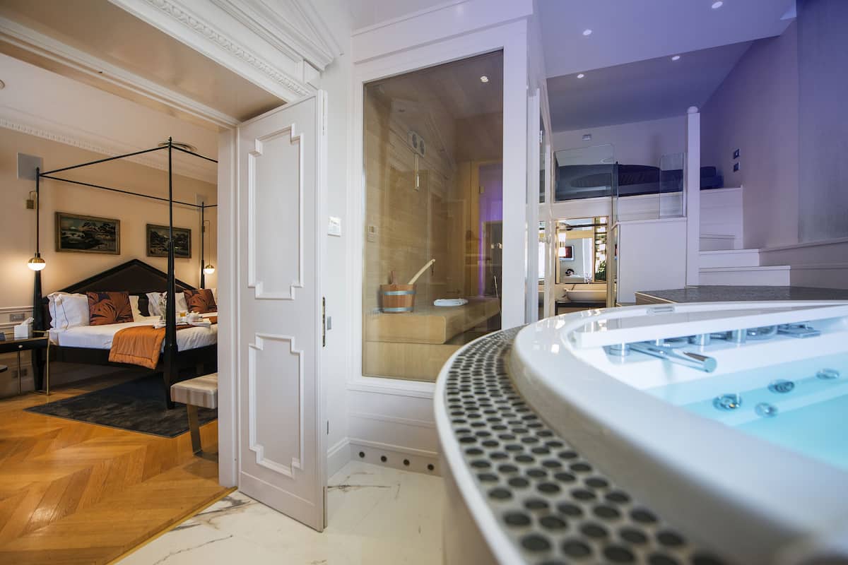 A modern hotel room with a large bathtub, a bed with a canopy, and a staircase leading to an upper level.