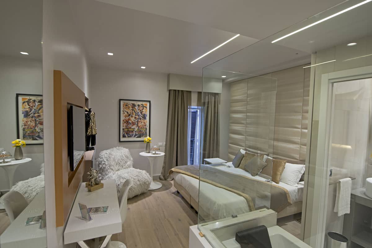 A modern hotel room with a large bed, a desk with a chair, a small table with a vase, and artwork on the walls.