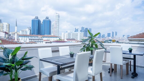 A rooftop terrace with a city skyline view, featuring a dining area with white chairs and a table, and potted plants.
