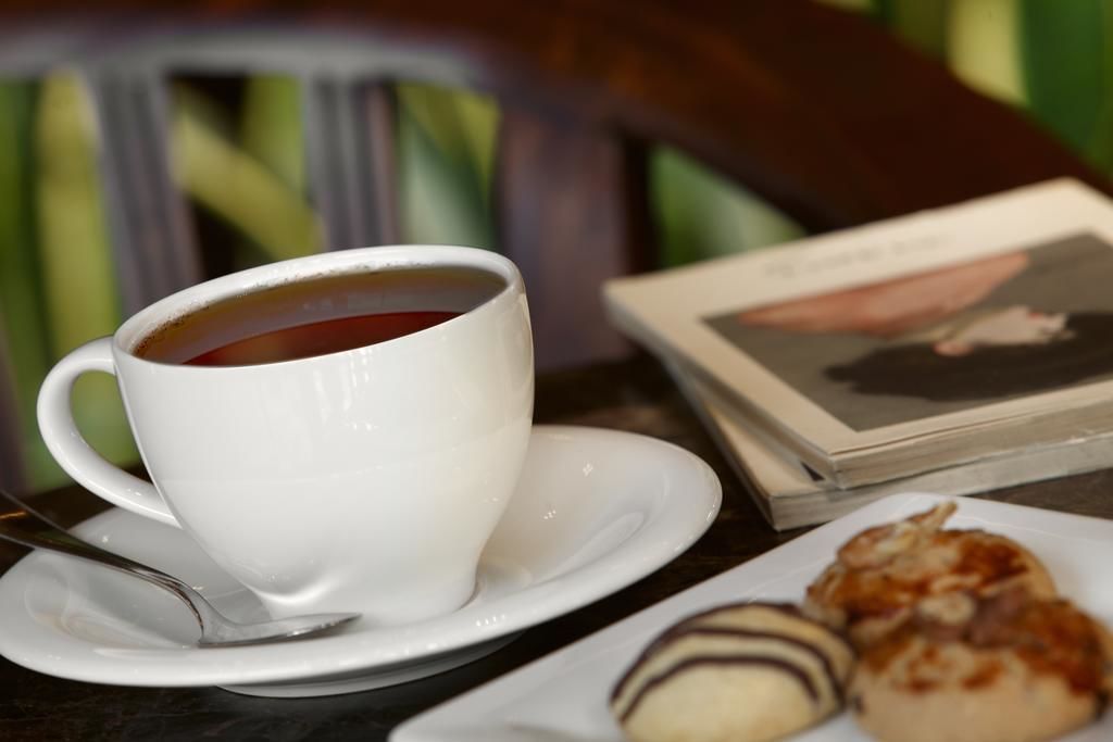 A white cup of tea on a saucer with a spoon, a plate with pastries, and a book on a table.