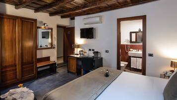 Ein Hotelzimmer mit Holzbalkendecke, einem Bett, einer Bank, einem Schreibtisch mit Lampe, einem Fernseher und einem Badezimmer mit Spiegel und Waschbecken.
