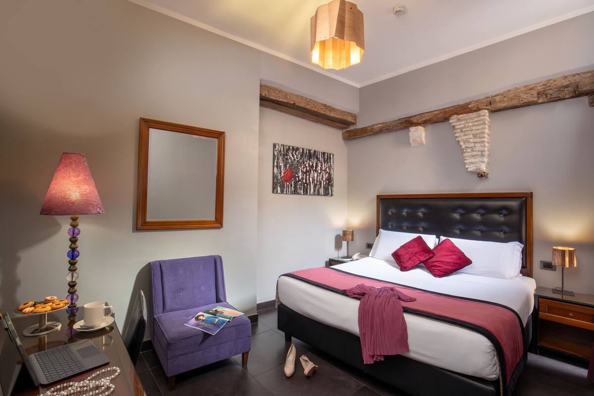 A hotel room with a bed, a chair, a desk with a laptop, a lamp, and a painting on the wall.