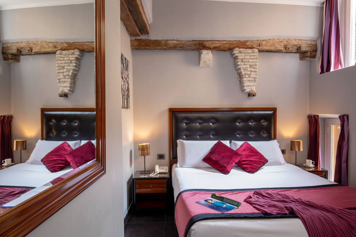 A hotel room with two beds, wooden beams, a mirror, bedside tables with lamps, and a window with curtains.