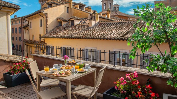 Una terraza con una mesa lista para el desayuno, rodeada de macetas con plantas y vistas a un paisaje urbano con edificios históricos.
