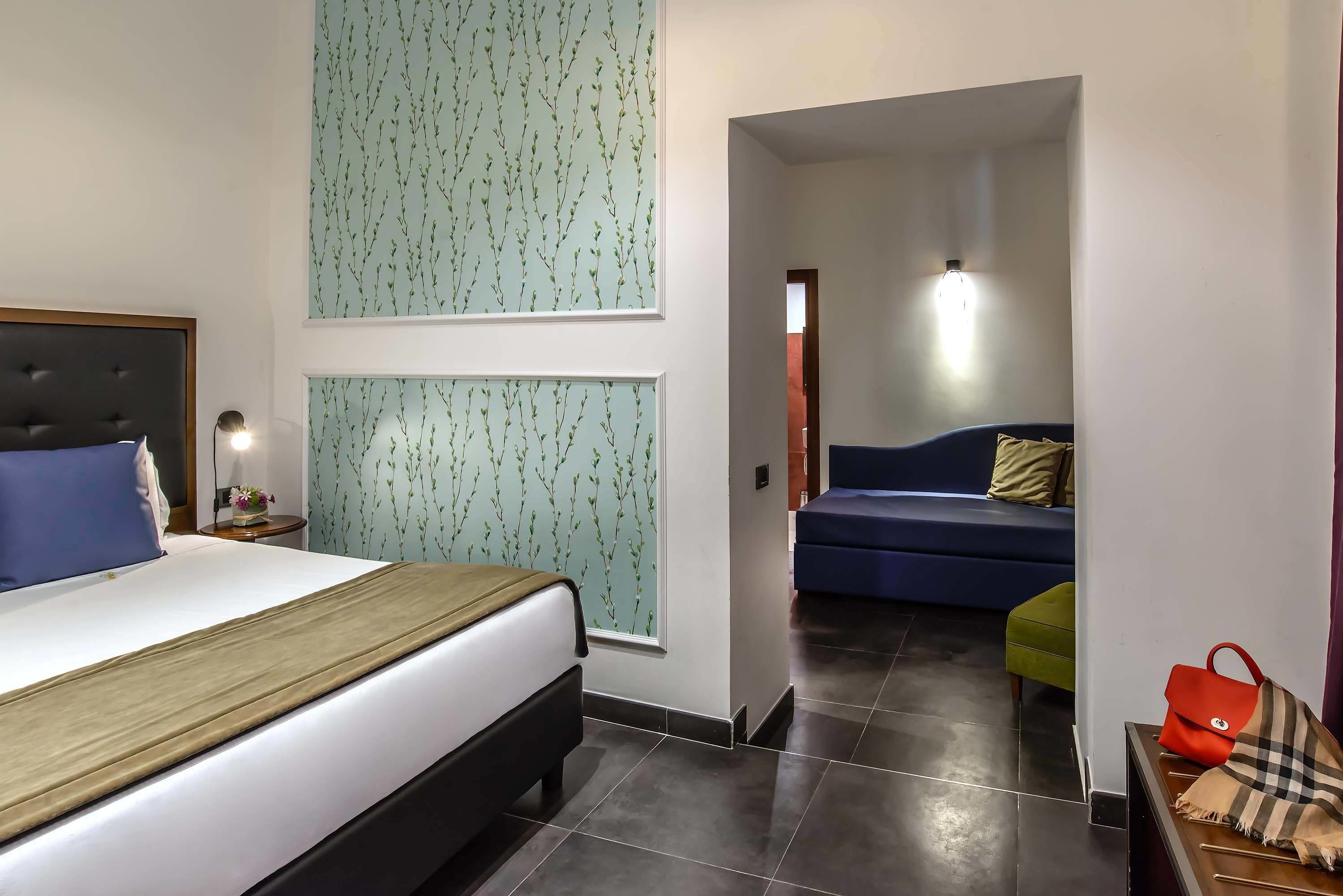 Een hotelkamer met een bed, nachtkastje, lamp, raam met uitzicht, blauwe bank en rode handtas op een tafel.