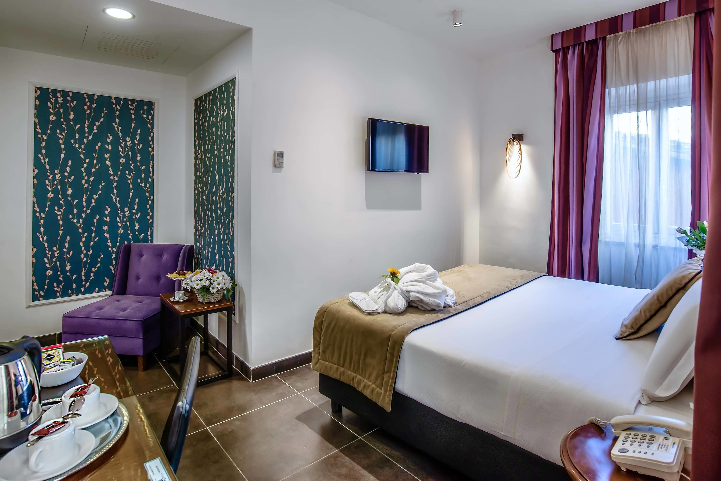 A hotel room with a bed, a chair, a table with a vase of flowers, a TV mounted on the wall, and a phone on the bedside table.
