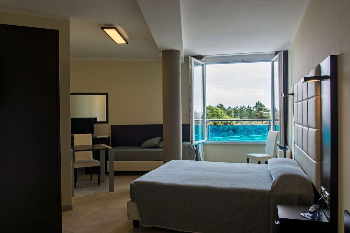 A modern hotel room with a large bed, a desk, a chair, and a balcony with a view of the sea.