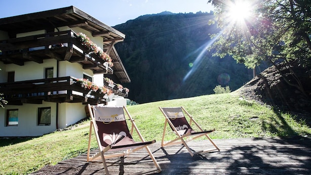 A chalet with wooden balconies and flower boxes, two wooden deck chairs, and a mountainous backdrop.