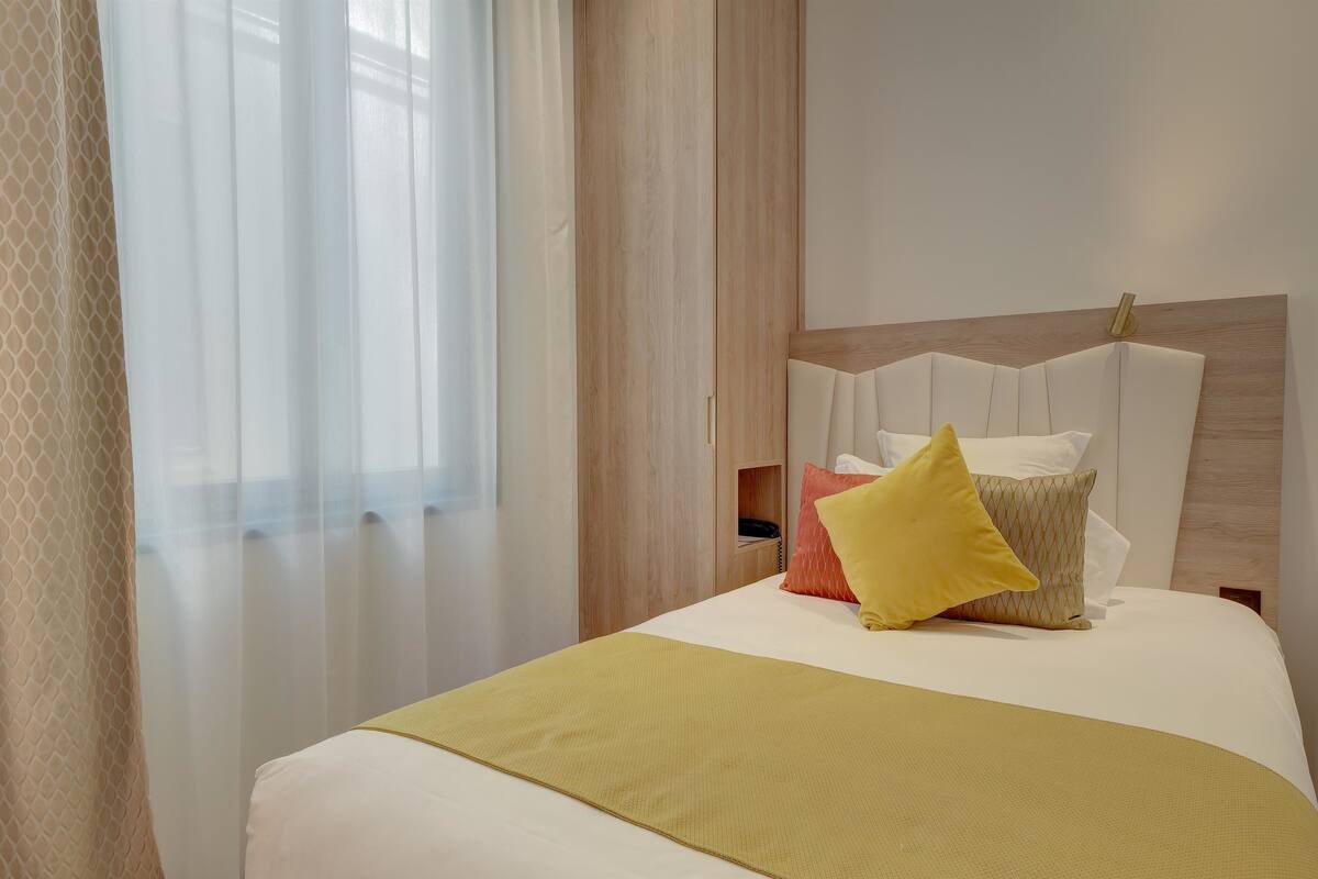 A neatly made bed with a yellow throw blanket, a wooden headboard, and a bedside table with a phone and a red pillow.