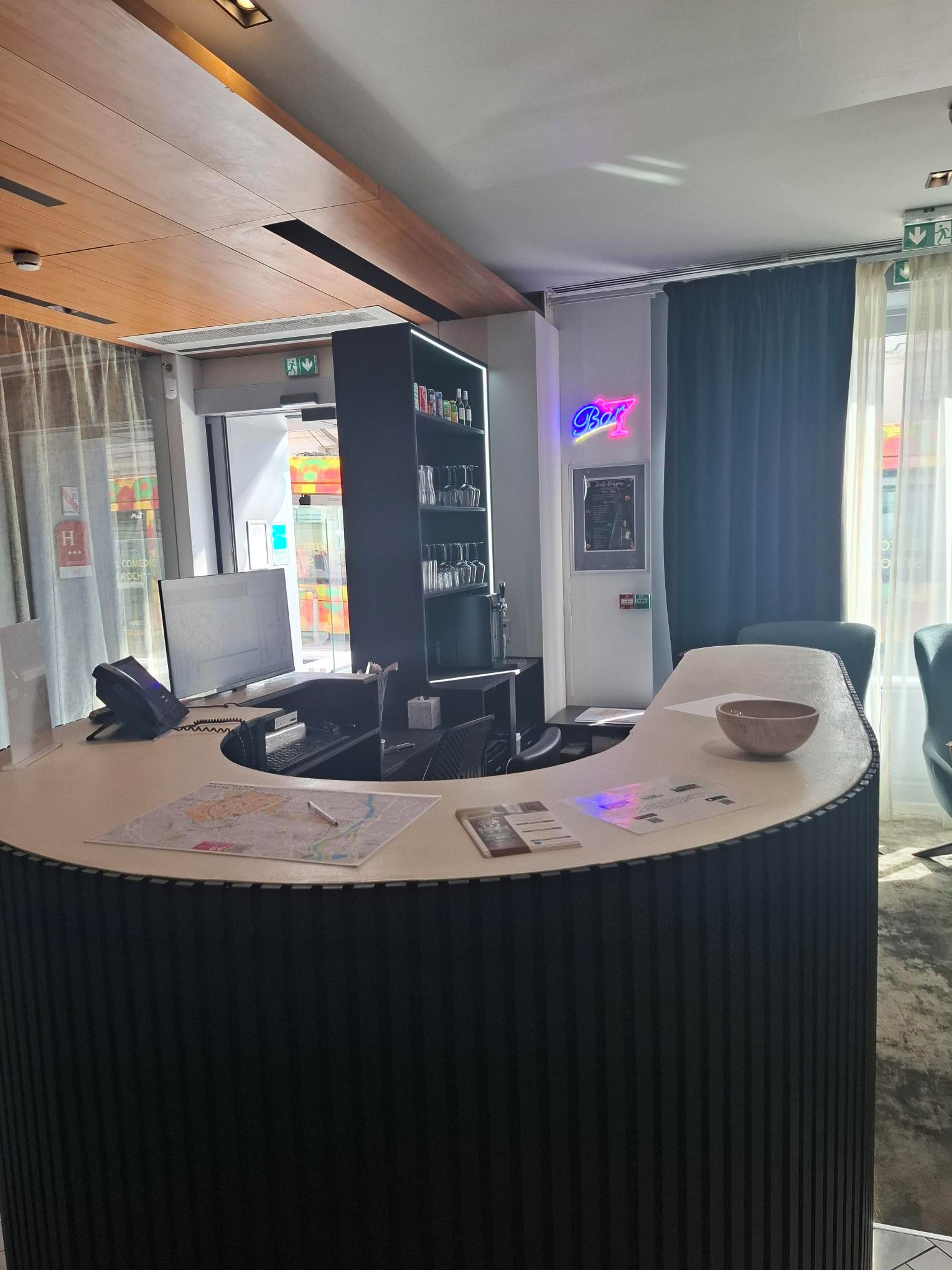Un comptoir d’accueil avec un ordinateur, un téléphone et une carte. On y trouve également une bibliothèque contenant divers objets et un panneau lumineux au néon.