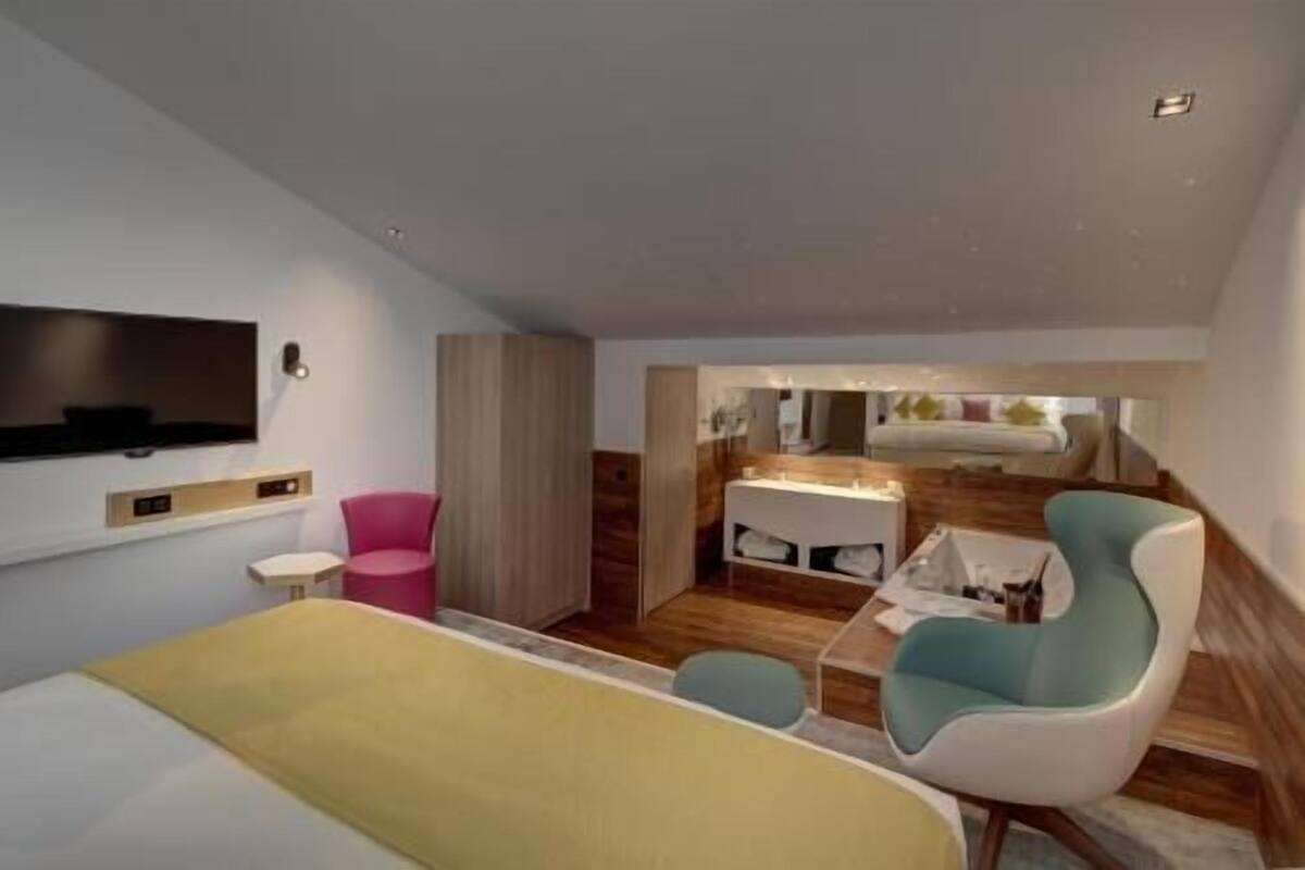 A modern hotel room with a bed, a TV, a seating area with a chair, and a small table.
