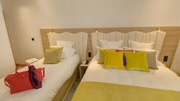 A hotel room with two beds, each with a wooden headboard, white bedding, and a variety of pillows. There is a red handbag on the left bed and a small bedside table with a phone and a notepad.
