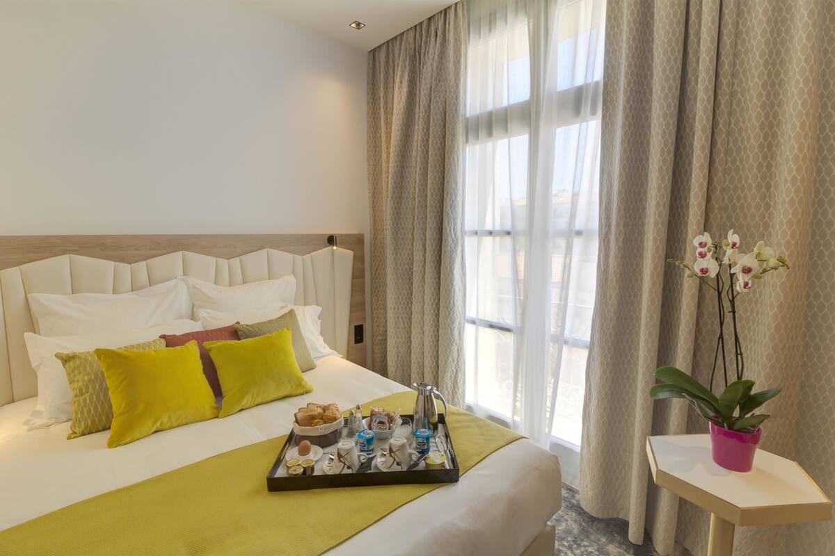 A hotel room with a neatly made bed, a tray of breakfast items, and a potted plant.