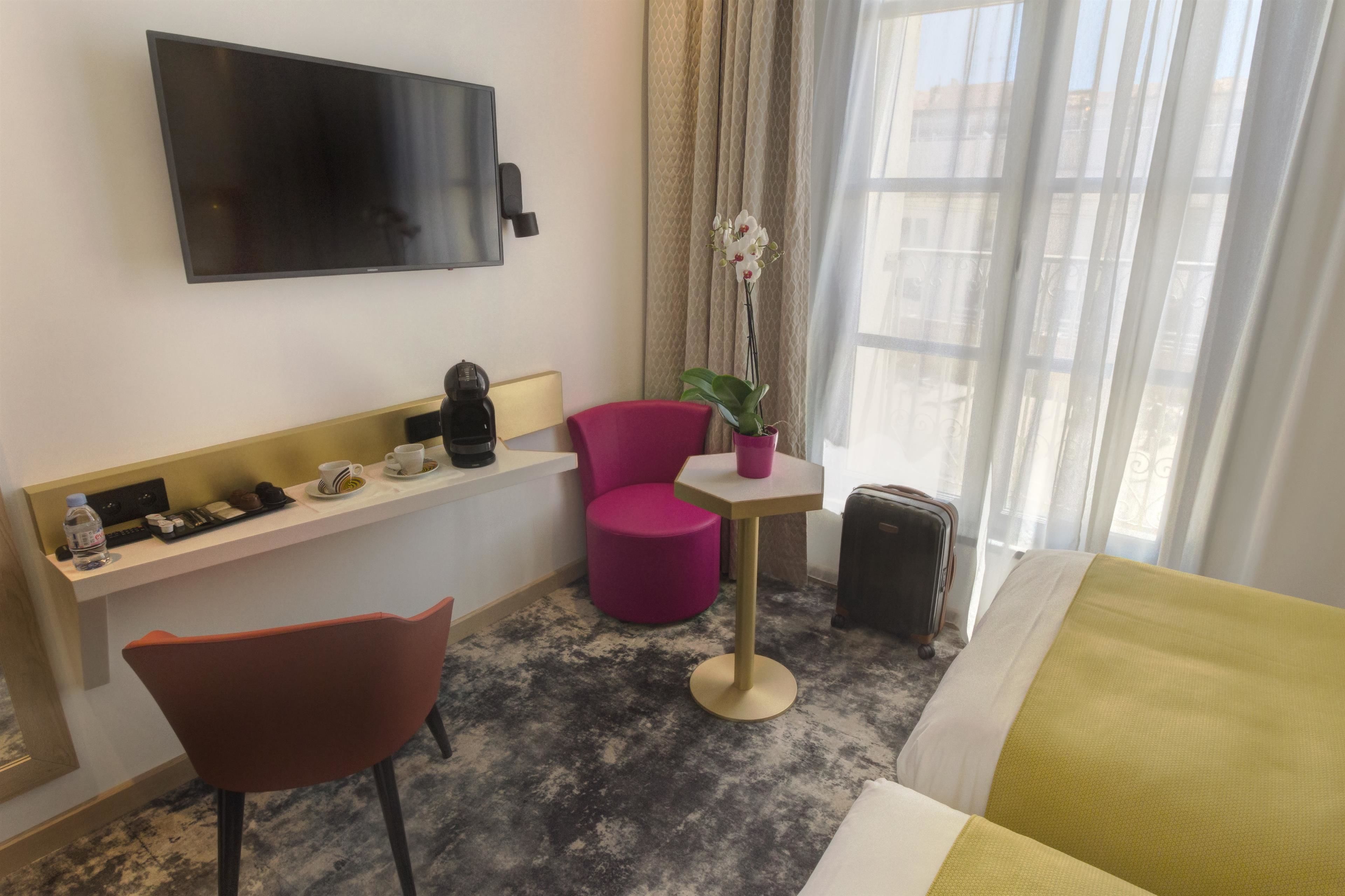 Habitación de hotel con cama, escritorio con silla, pequeña mesa con silla, planta en maceta y televisión montada en la pared.