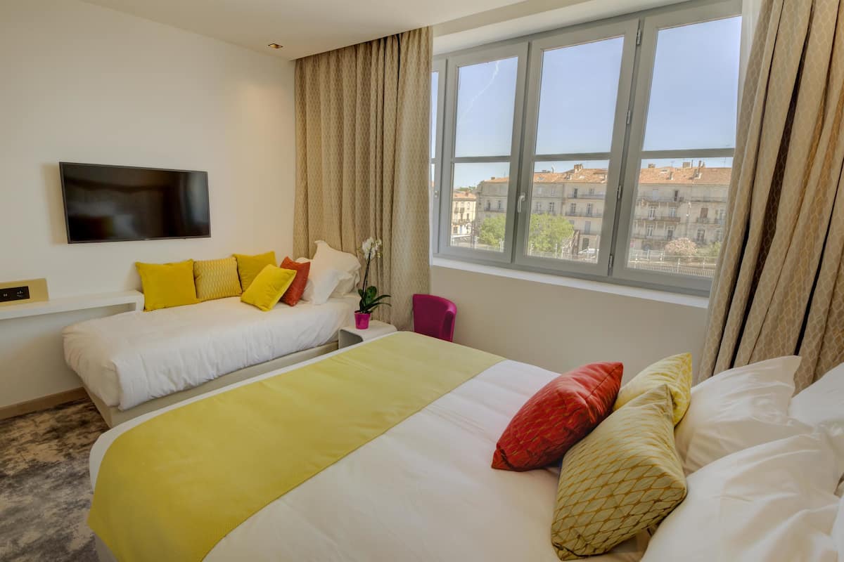 A hotel room with two beds, a TV, a window with curtains, and a view of buildings outside.