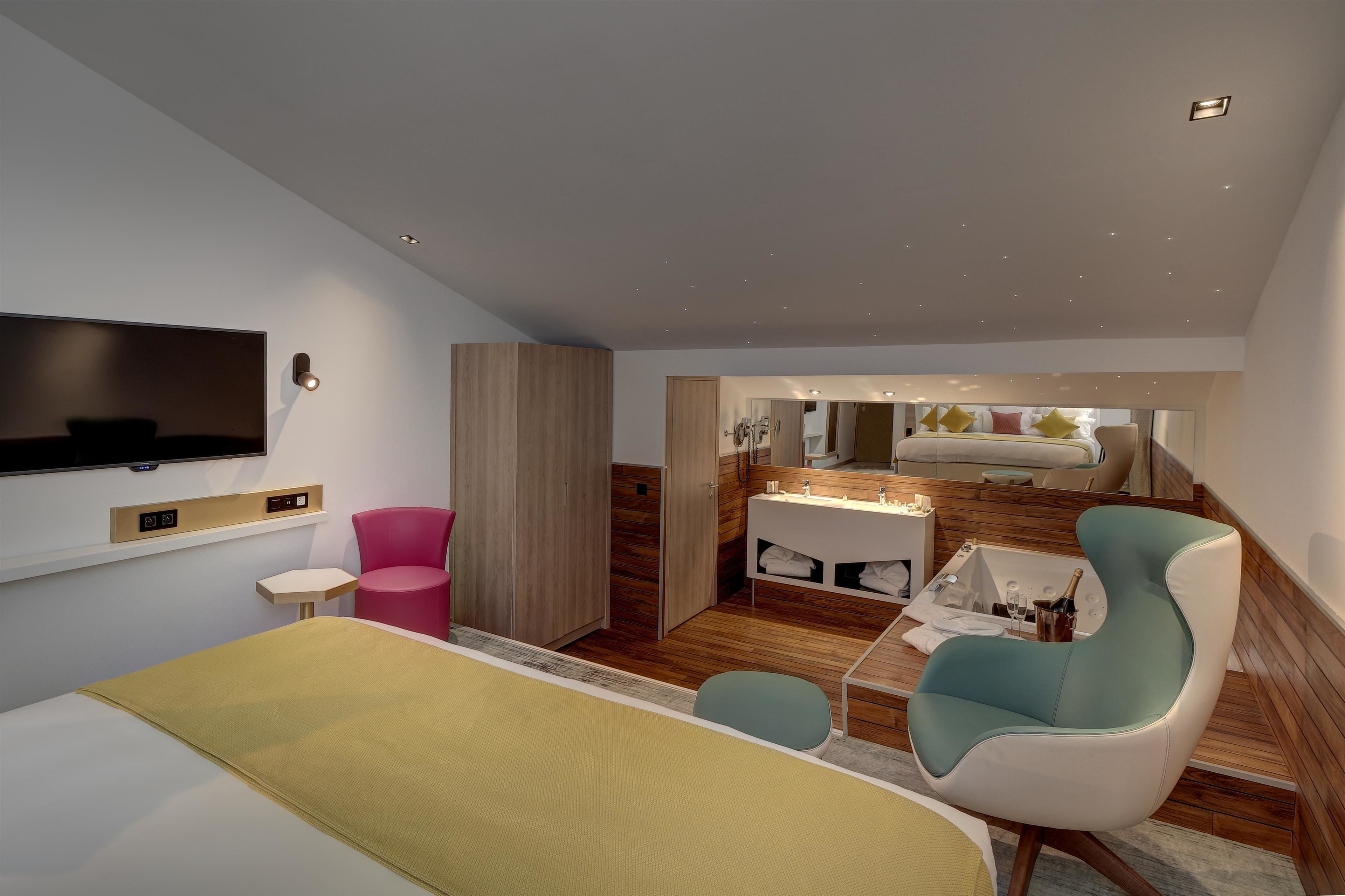 A modern hotel room with a large bed, a TV mounted on the wall, a small table, a pink chair, and a bathroom with a sink and mirror.