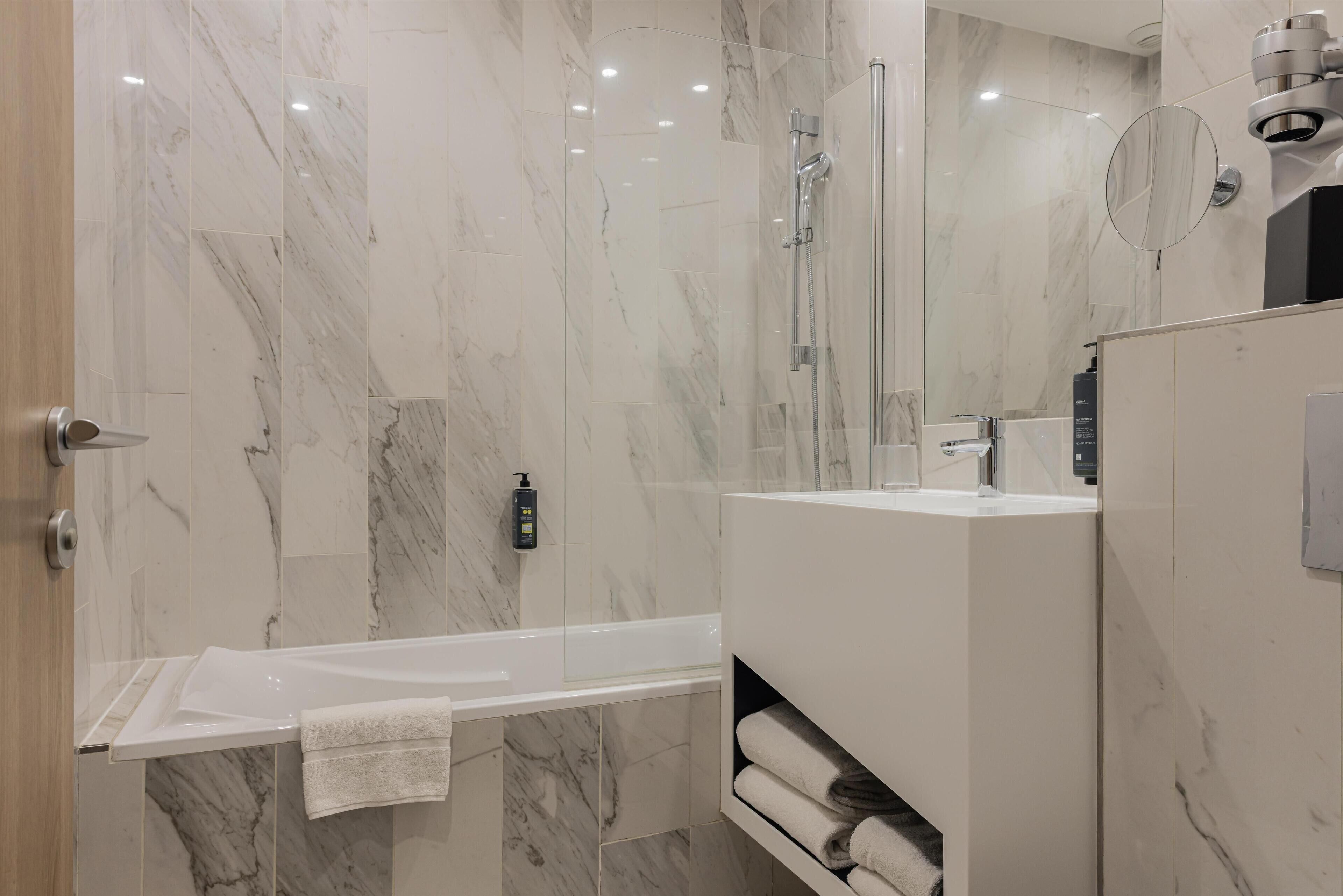 Een moderne badkamer met een marmeren vloer, een douche met glazen wand, een wit bad en een ingebouwde wastafel met spiegel.